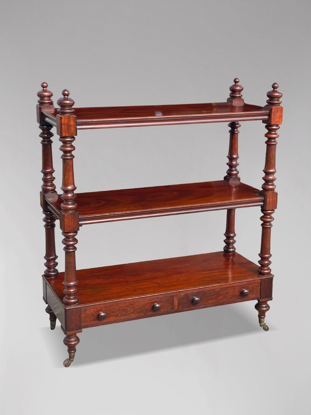 A nineteenth-century three-tiered mahogany dumb waiter with turned support columns, two drawers at the base, and brass castors.