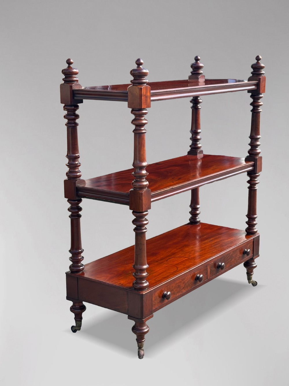 A nineteenth-century three-tiered mahogany dumb waiter with turned support columns, two drawers at the base, and brass castors. View IV