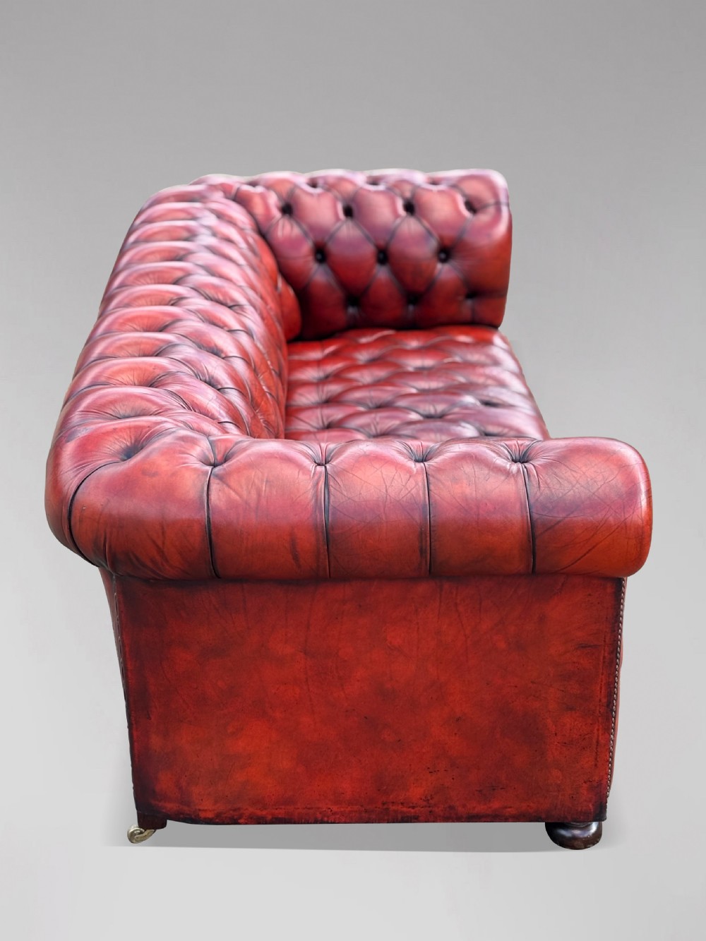 An antique three-seater Chesterfield sofa upholstered in deep red buttoned leather with rolled arms and brass studding. View V