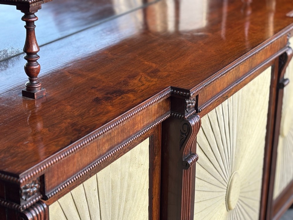 A Regency rosewood sideboard featuring three doors with sunburst pleated panels, a mirrored upper shelf supported by brass columns, and a pierced brass top gallery. View IX