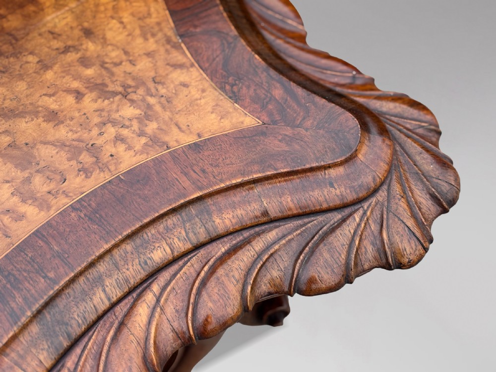 A 19th-century Victorian burr walnut centre table featuring a serpentine top with central floral marquetry and carved scroll legs on castors. View IV