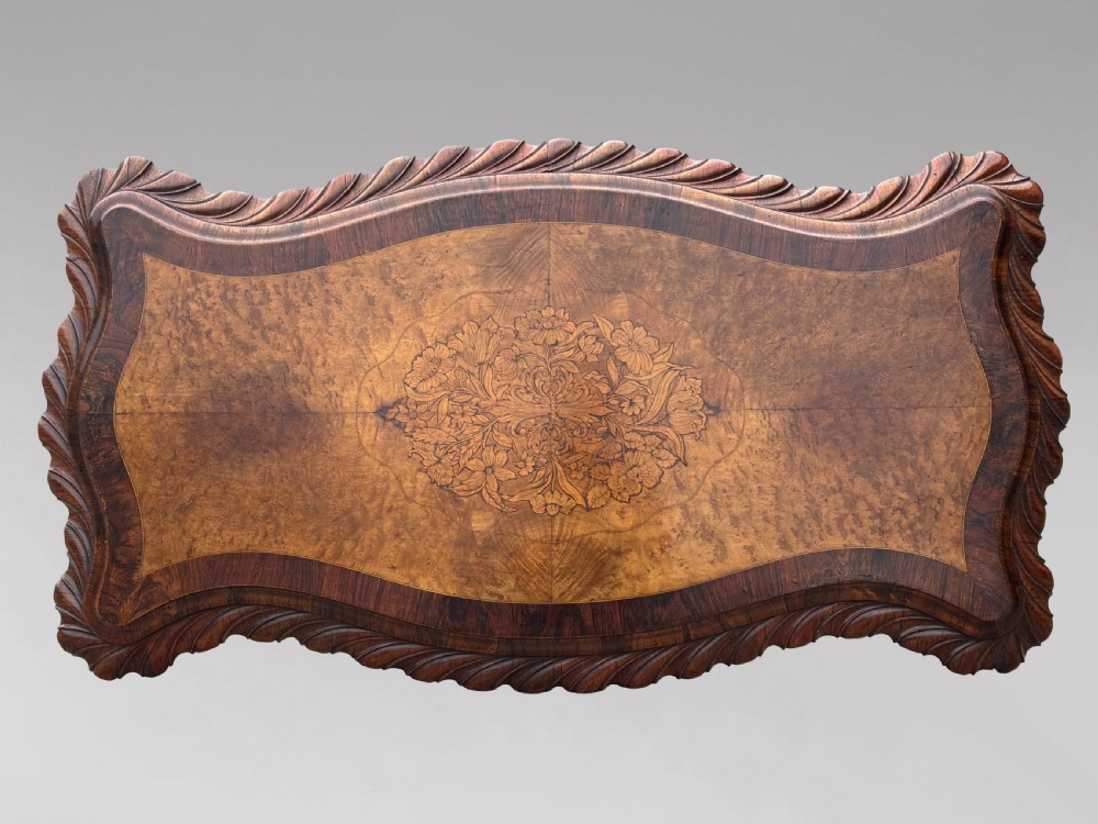 A 19th-century Victorian burr walnut centre table featuring a serpentine top with central floral marquetry and carved scroll legs on castors. View V