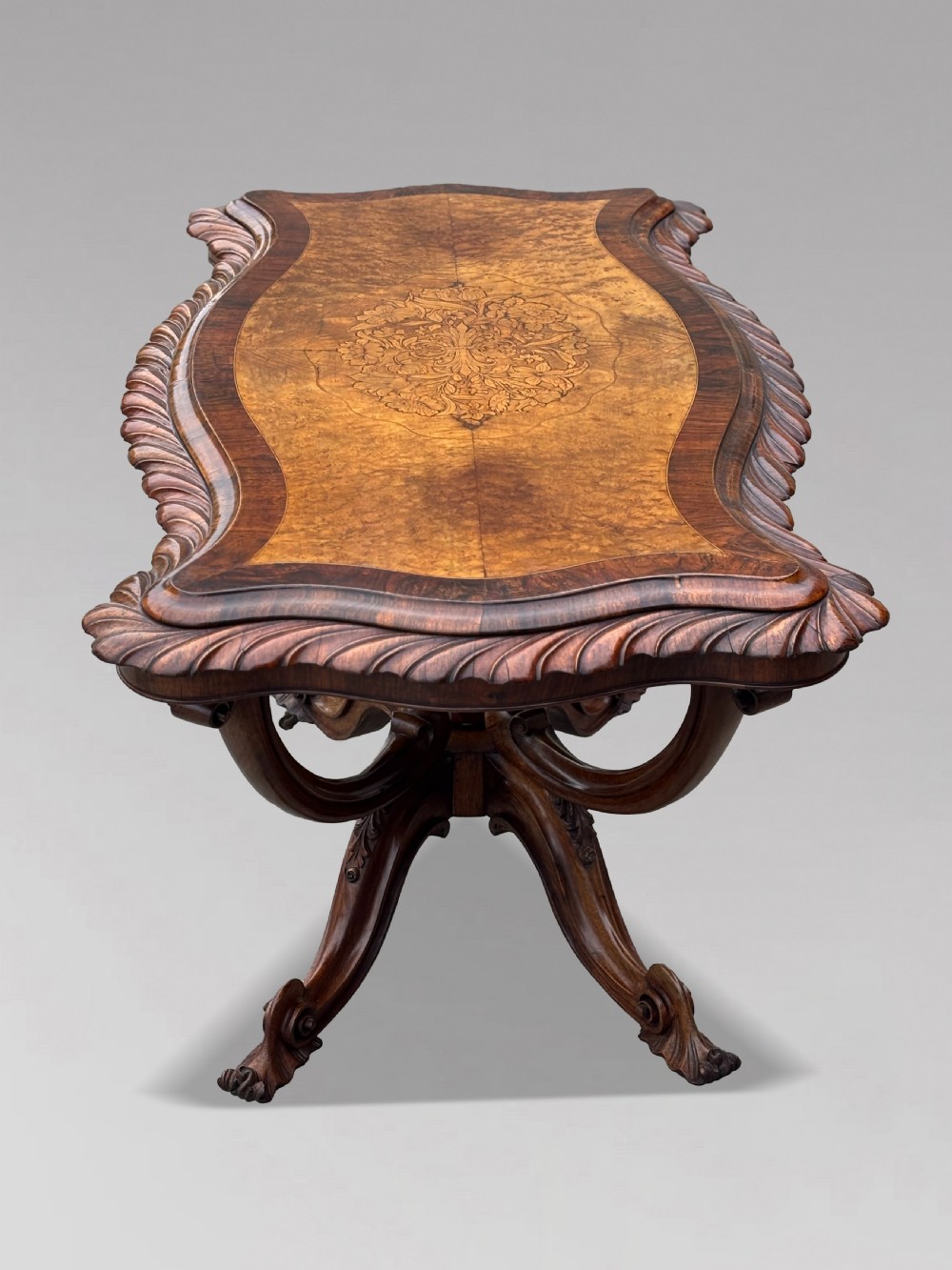 A 19th-century Victorian burr walnut centre table featuring a serpentine top with central floral marquetry and carved scroll legs on castors. View X