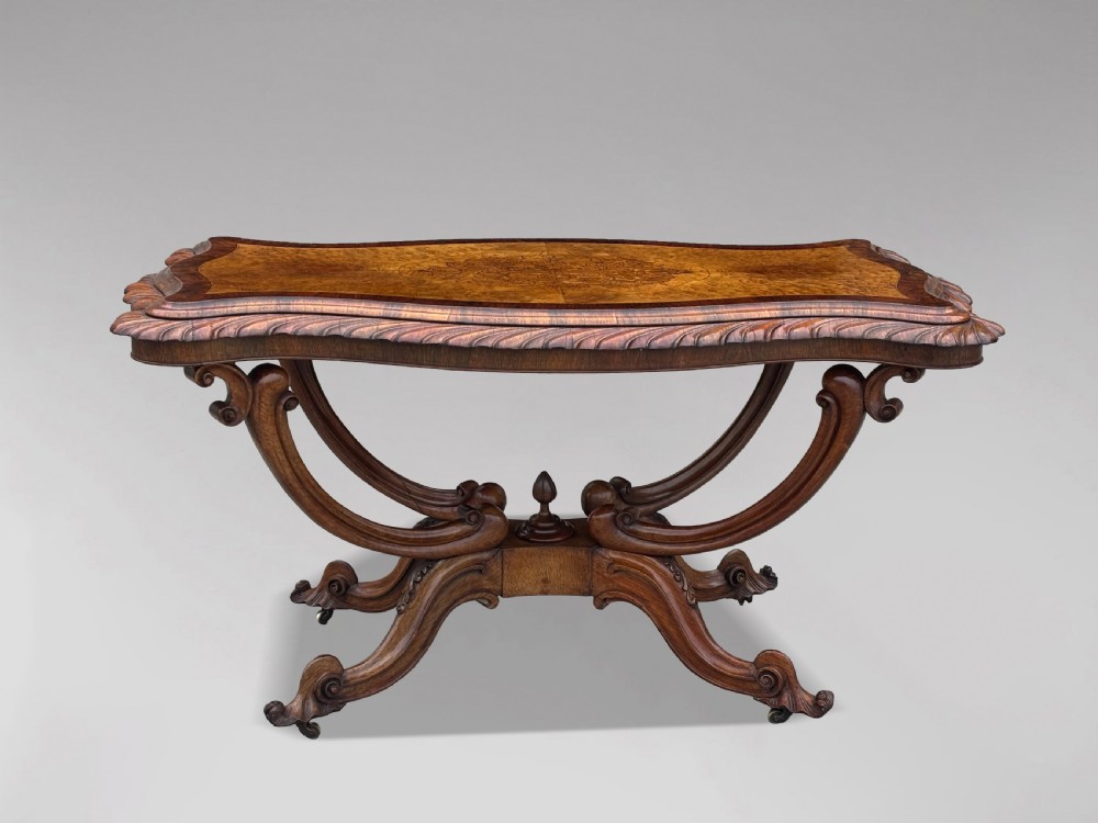 A 19th-century Victorian burr walnut centre table featuring a serpentine top with central floral marquetry and carved scroll legs on castors. View XI