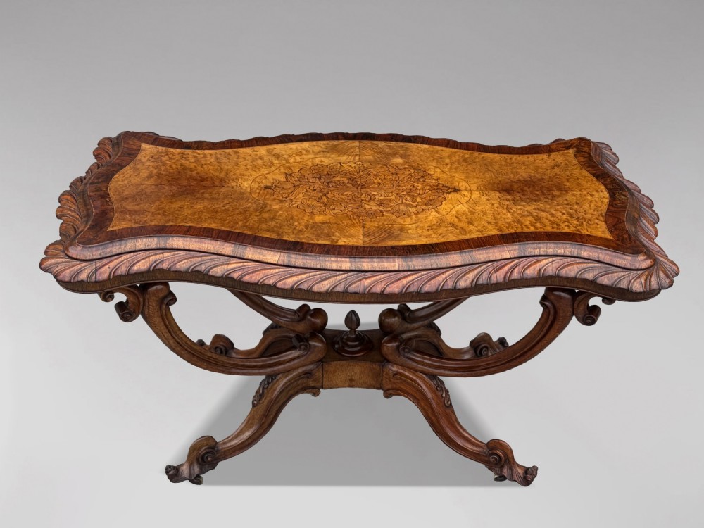 A 19th-century Victorian burr walnut centre table featuring a serpentine top with central floral marquetry and carved scroll legs on castors. View XII