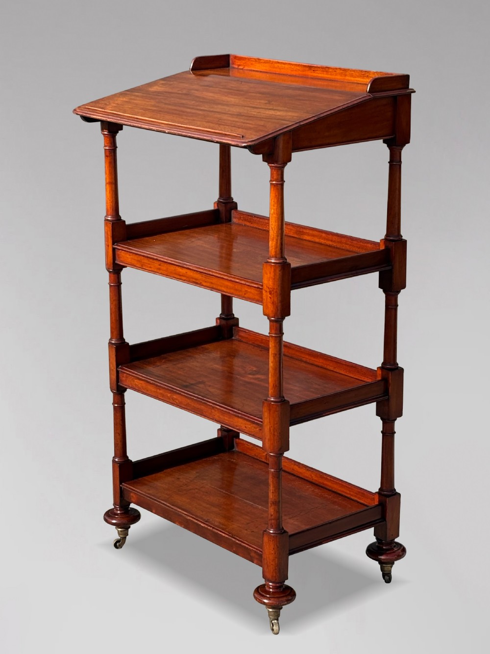 A four-tier George IV mahogany whatnot featuring a sloped top reading surface, turned wooden upright supports, and brass castors at the feet.