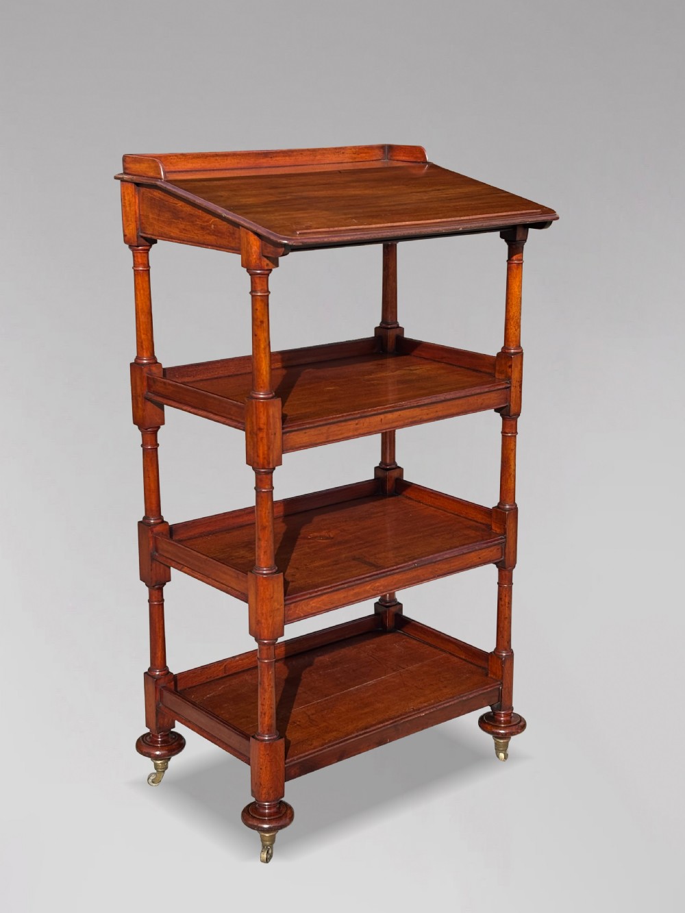 A four-tier George IV mahogany whatnot featuring a sloped top reading surface, turned wooden upright supports, and brass castors at the feet. View II