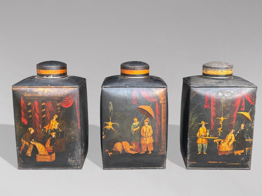 A set of three black rectangular antique toleware tea canisters hand-painted with detailed Chinoiserie figures and red drapery scenes.