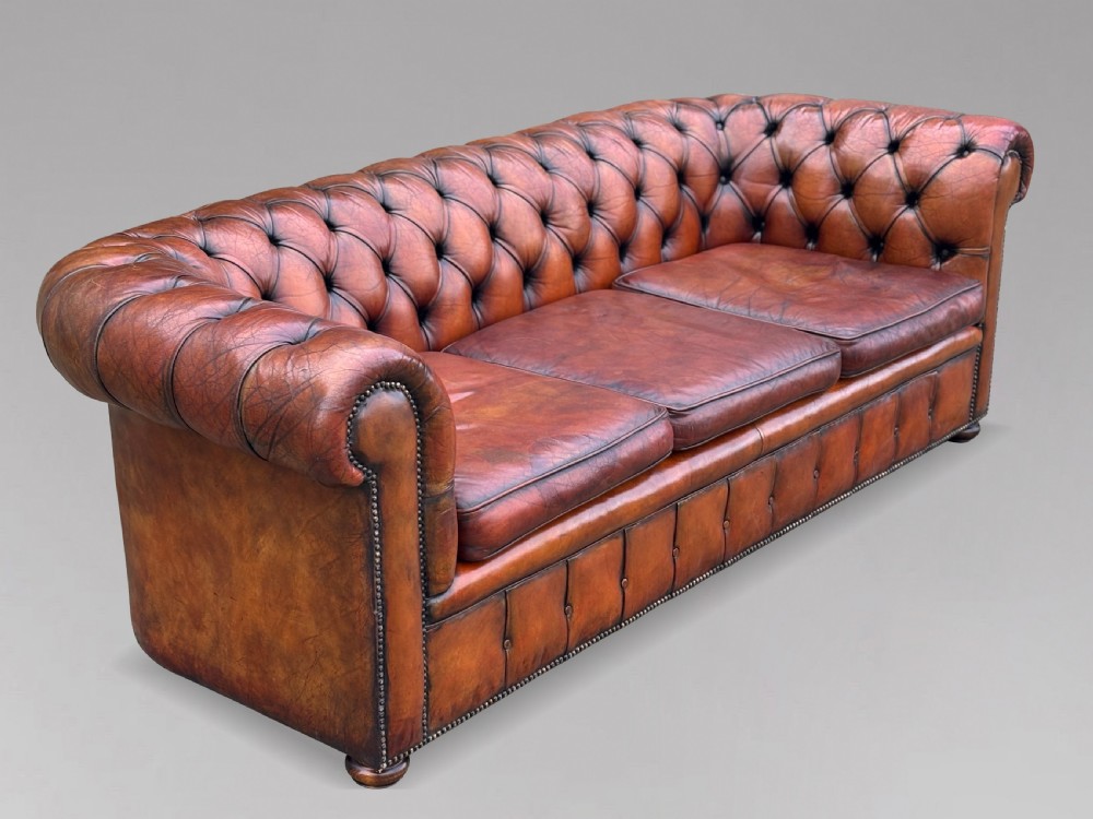 A three-seater brown leather Chesterfield sofa featuring deep button-tufted upholstery, rolled arms, brass studding, and three removable seat cushions. View IV