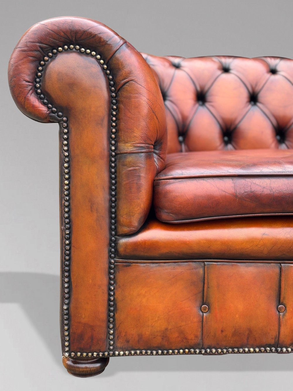 A three-seater brown leather Chesterfield sofa featuring deep button-tufted upholstery, rolled arms, brass studding, and three removable seat cushions. View VI