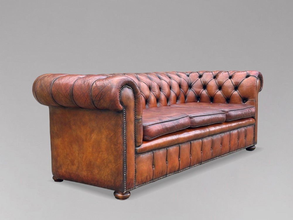 A three-seater brown leather Chesterfield sofa featuring deep button-tufted upholstery, rolled arms, brass studding, and three removable seat cushions. View IX