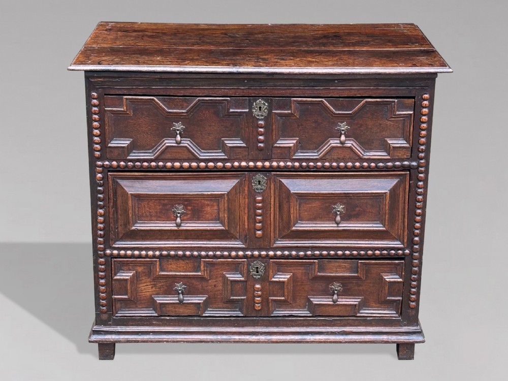 A 17th-century dark oak chest of drawers featuring three drawers decorated with raised geometric mitred mouldings, beaded trim details, and central brass drop handles.