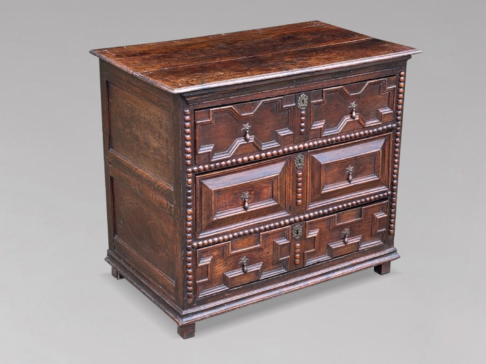 A 17th-century dark oak chest of drawers featuring three drawers decorated with raised geometric mitred mouldings, beaded trim details, and central brass drop handles. View II