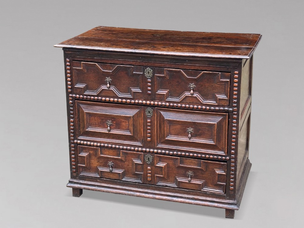 A 17th-century dark oak chest of drawers featuring three drawers decorated with raised geometric mitred mouldings, beaded trim details, and central brass drop handles. View IV