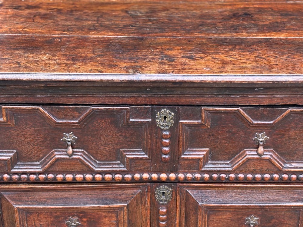 A 17th-century dark oak chest of drawers featuring three drawers decorated with raised geometric mitred mouldings, beaded trim details, and central brass drop handles. View V
