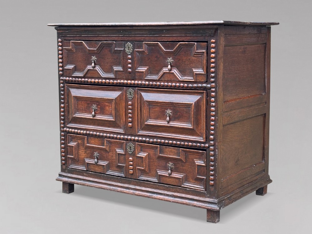 A 17th-century dark oak chest of drawers featuring three drawers decorated with raised geometric mitred mouldings, beaded trim details, and central brass drop handles. View VI