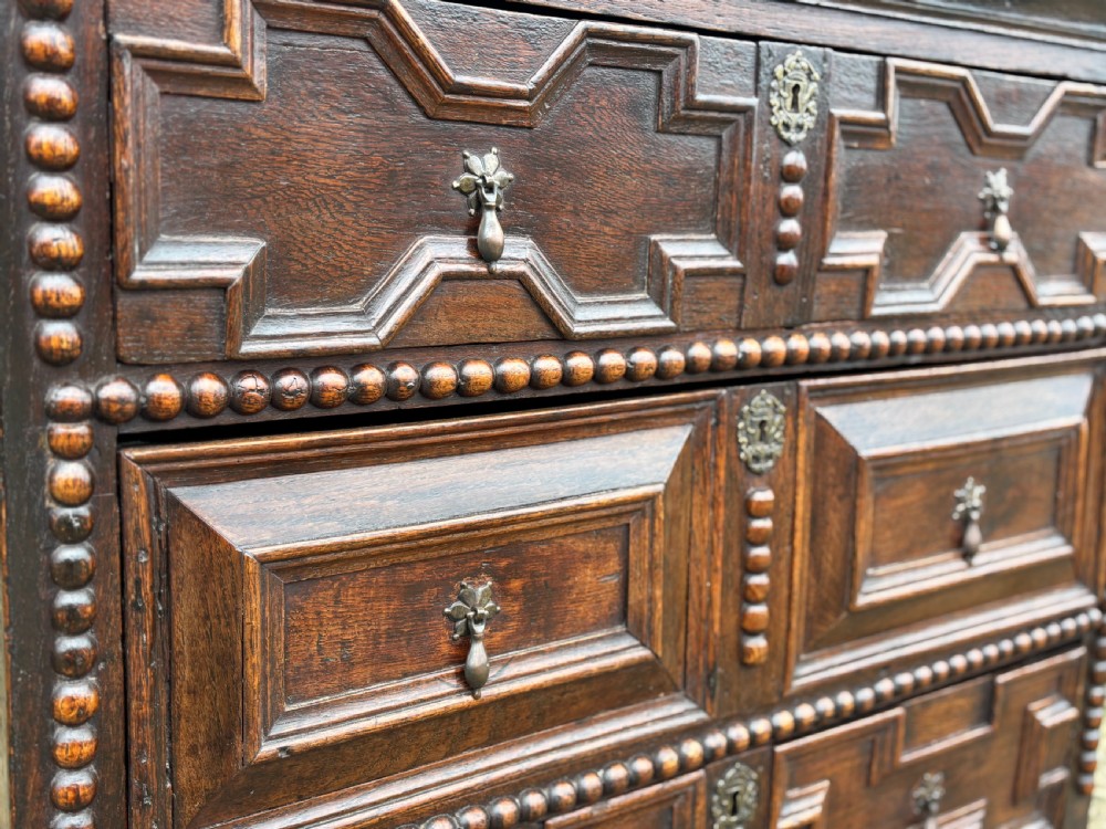 A 17th-century dark oak chest of drawers featuring three drawers decorated with raised geometric mitred mouldings, beaded trim details, and central brass drop handles. View VII
