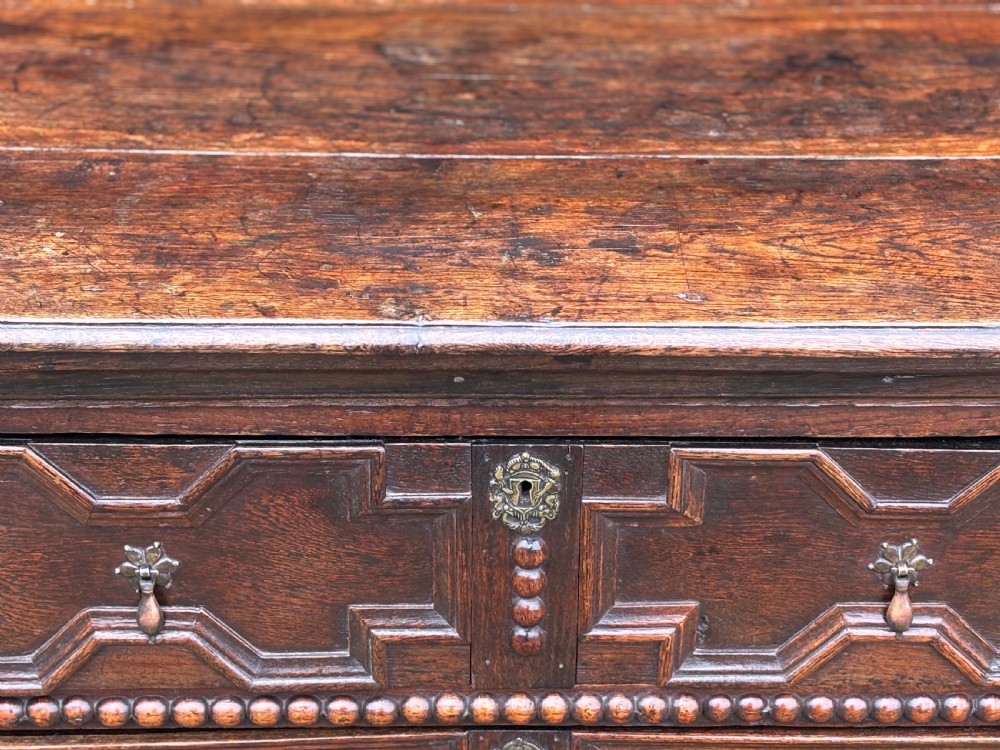 A 17th-century dark oak chest of drawers featuring three drawers decorated with raised geometric mitred mouldings, beaded trim details, and central brass drop handles. View IX