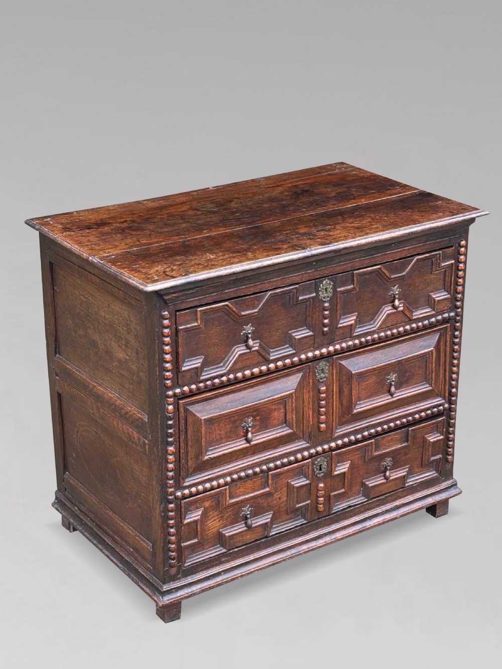 A 17th-century dark oak chest of drawers featuring three drawers decorated with raised geometric mitred mouldings, beaded trim details, and central brass drop handles. View XI