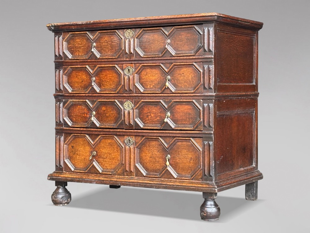 A seventeenth-century Charles II oak chest of drawers featuring four geometric panelled drawers with brass teardrop handles, resting on turned bun feet.