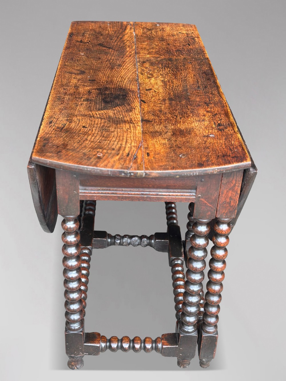 A 17th-century Charles II dark oak gateleg table featuring an oval drop-leaf top supported by bobbin-turned legs and stretchers. View V
