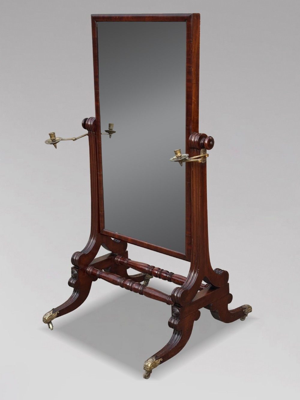A 19th-century William IV mahogany cheval mirror featuring a rectangular swivel glass plate, dual brass candle sconces, turned stretchers, and splayed legs with brass claw feet on castors.