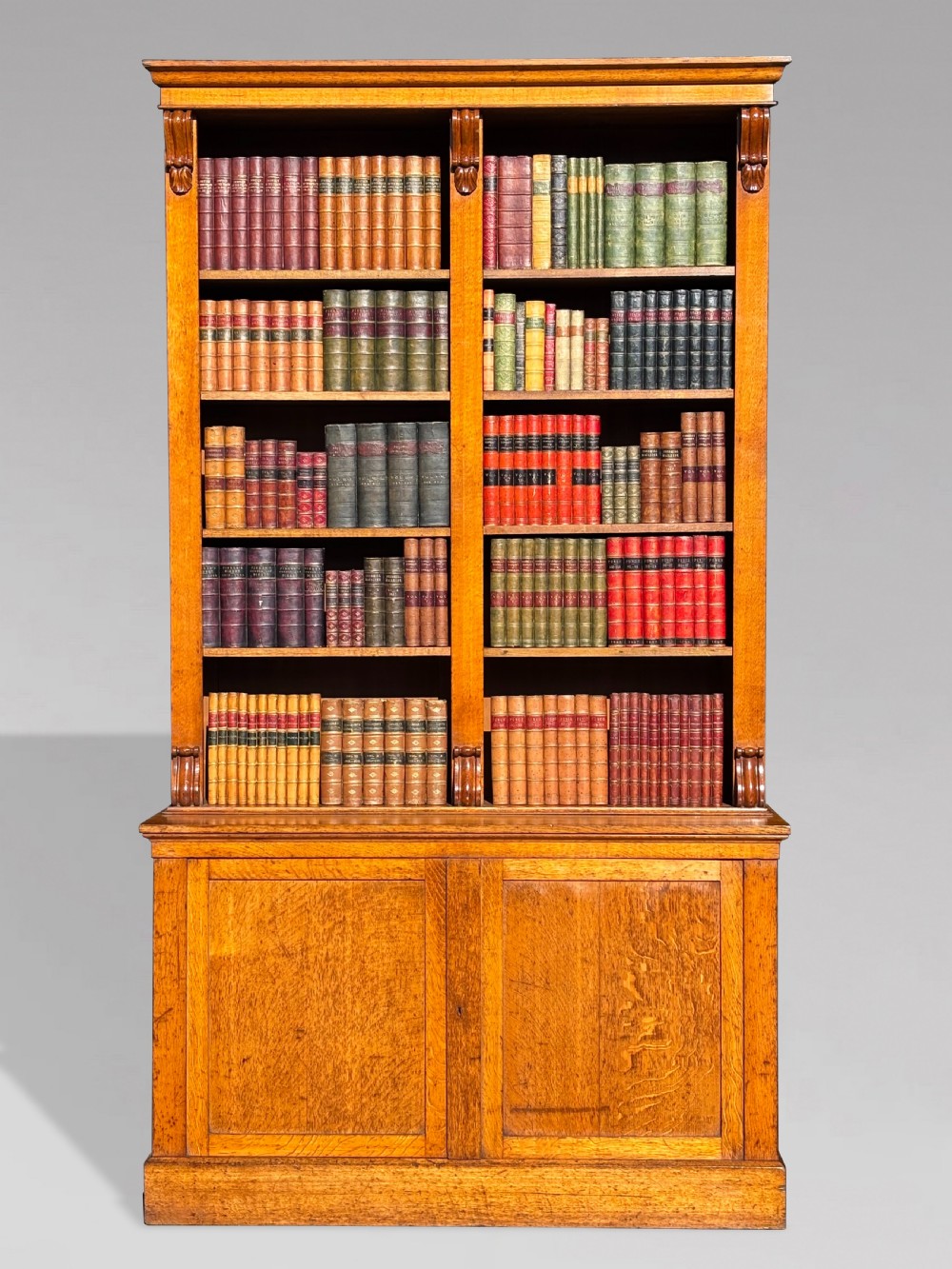 A two-section 19th-century oak open bookcase featuring carved corbels flanking the upper shelves and a double-panelled cupboard base.