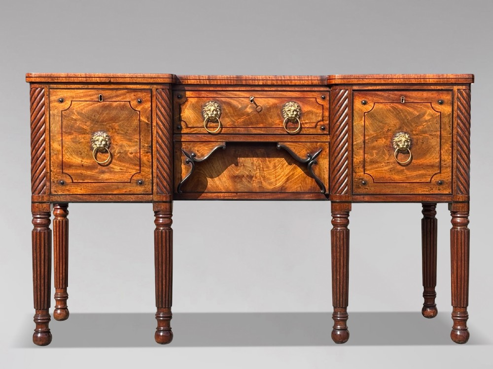 A 19th-century George IV flame mahogany breakfront sideboard featuring ebony stringing, lion-mask brass handles, spiral pilasters, and fluted legs.