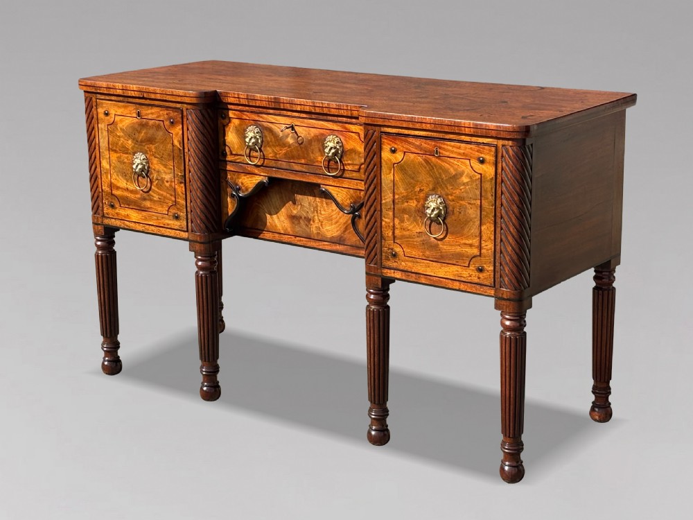 A 19th-century George IV flame mahogany breakfront sideboard featuring ebony stringing, lion-mask brass handles, spiral pilasters, and fluted legs. View II