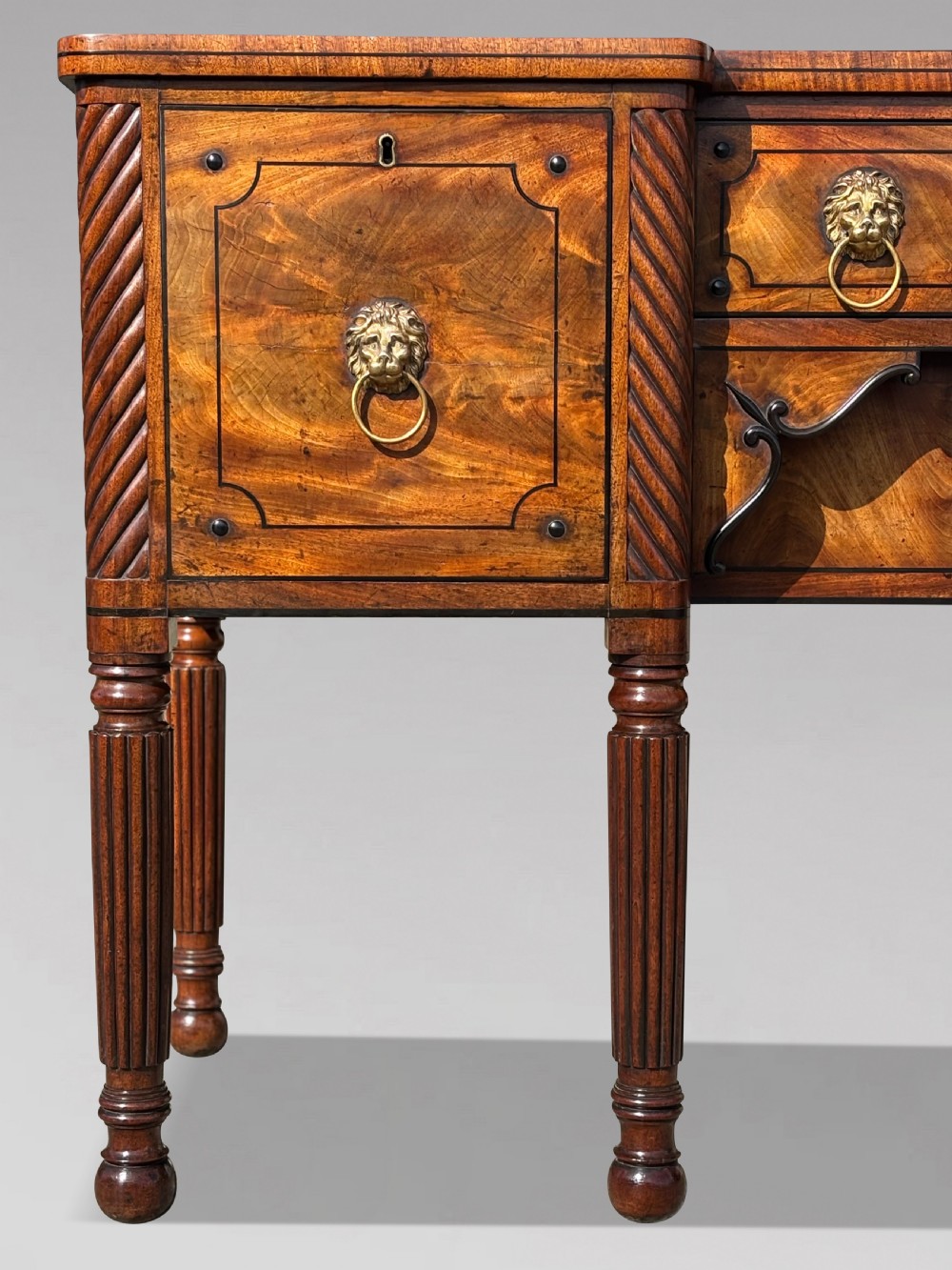 A 19th-century George IV flame mahogany breakfront sideboard featuring ebony stringing, lion-mask brass handles, spiral pilasters, and fluted legs. View IV
