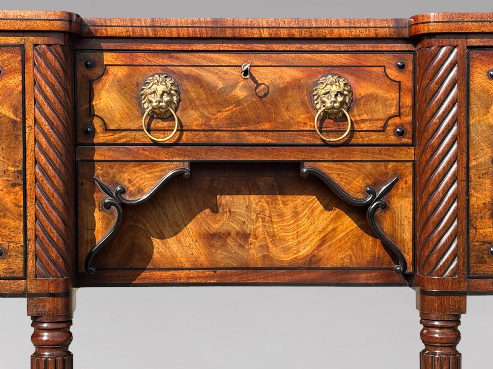 A 19th-century George IV flame mahogany breakfront sideboard featuring ebony stringing, lion-mask brass handles, spiral pilasters, and fluted legs. View V