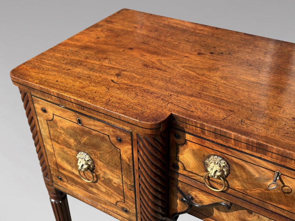 A 19th-century George IV flame mahogany breakfront sideboard featuring ebony stringing, lion-mask brass handles, spiral pilasters, and fluted legs. View VI