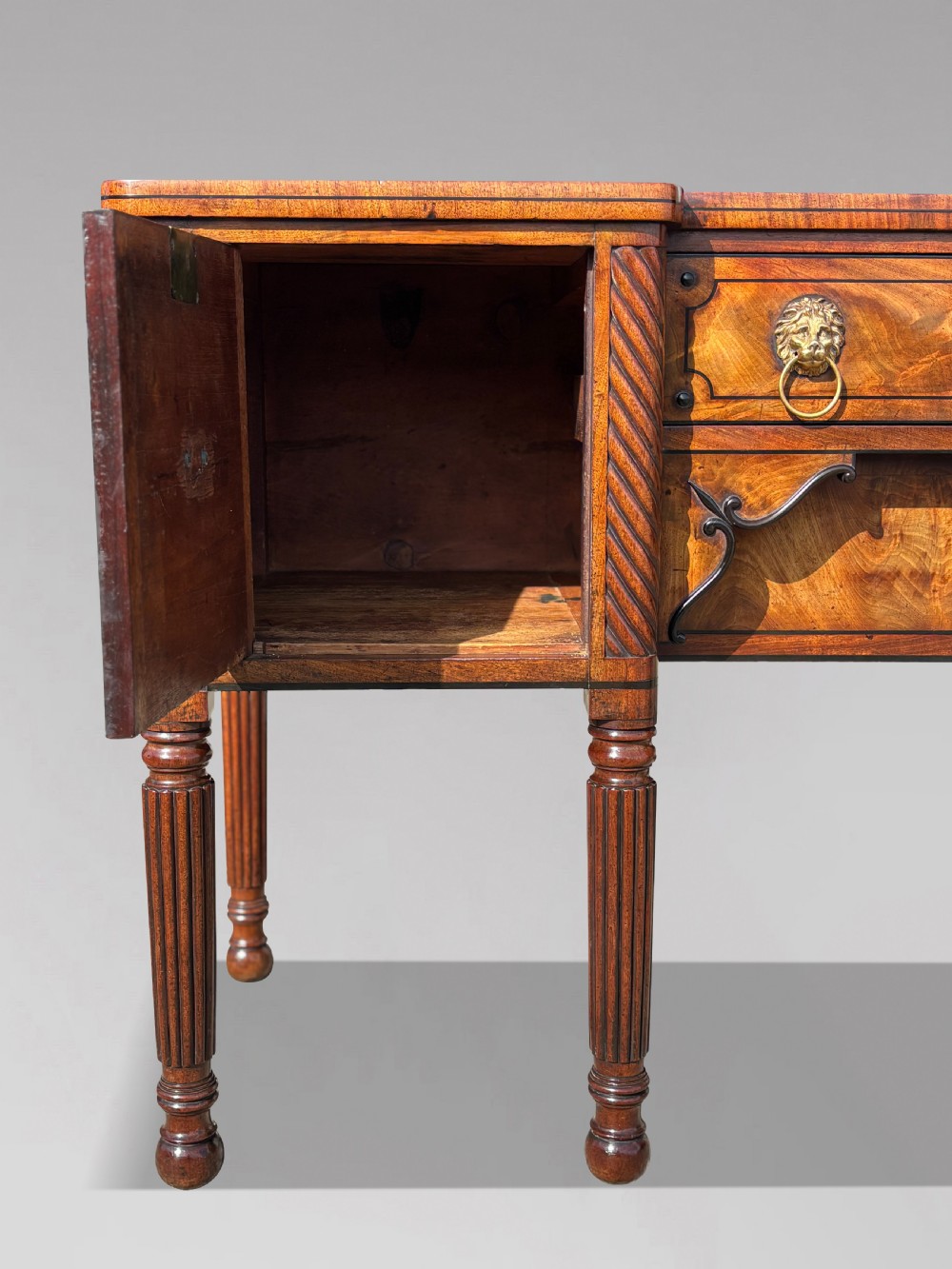 A 19th-century George IV flame mahogany breakfront sideboard featuring ebony stringing, lion-mask brass handles, spiral pilasters, and fluted legs. View XII