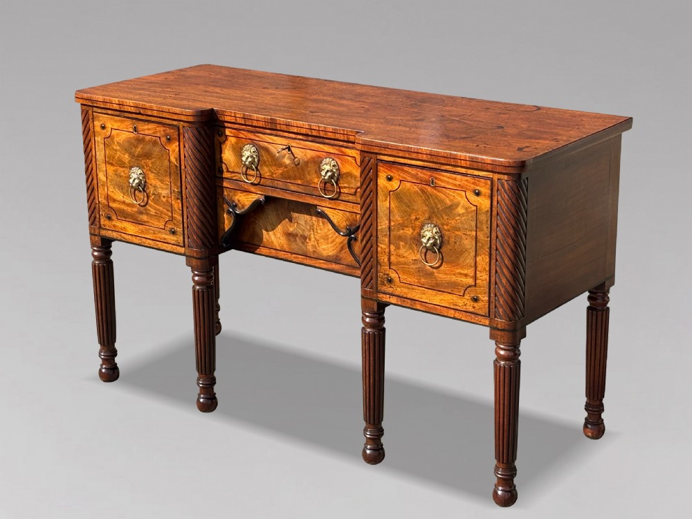 A 19th-century George IV flame mahogany breakfront sideboard featuring ebony stringing, lion-mask brass handles, spiral pilasters, and fluted legs. View XIII