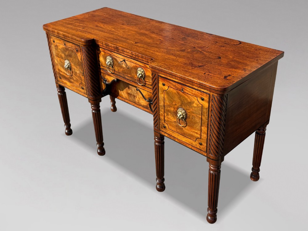 A 19th-century George IV flame mahogany breakfront sideboard featuring ebony stringing, lion-mask brass handles, spiral pilasters, and fluted legs. View XIV