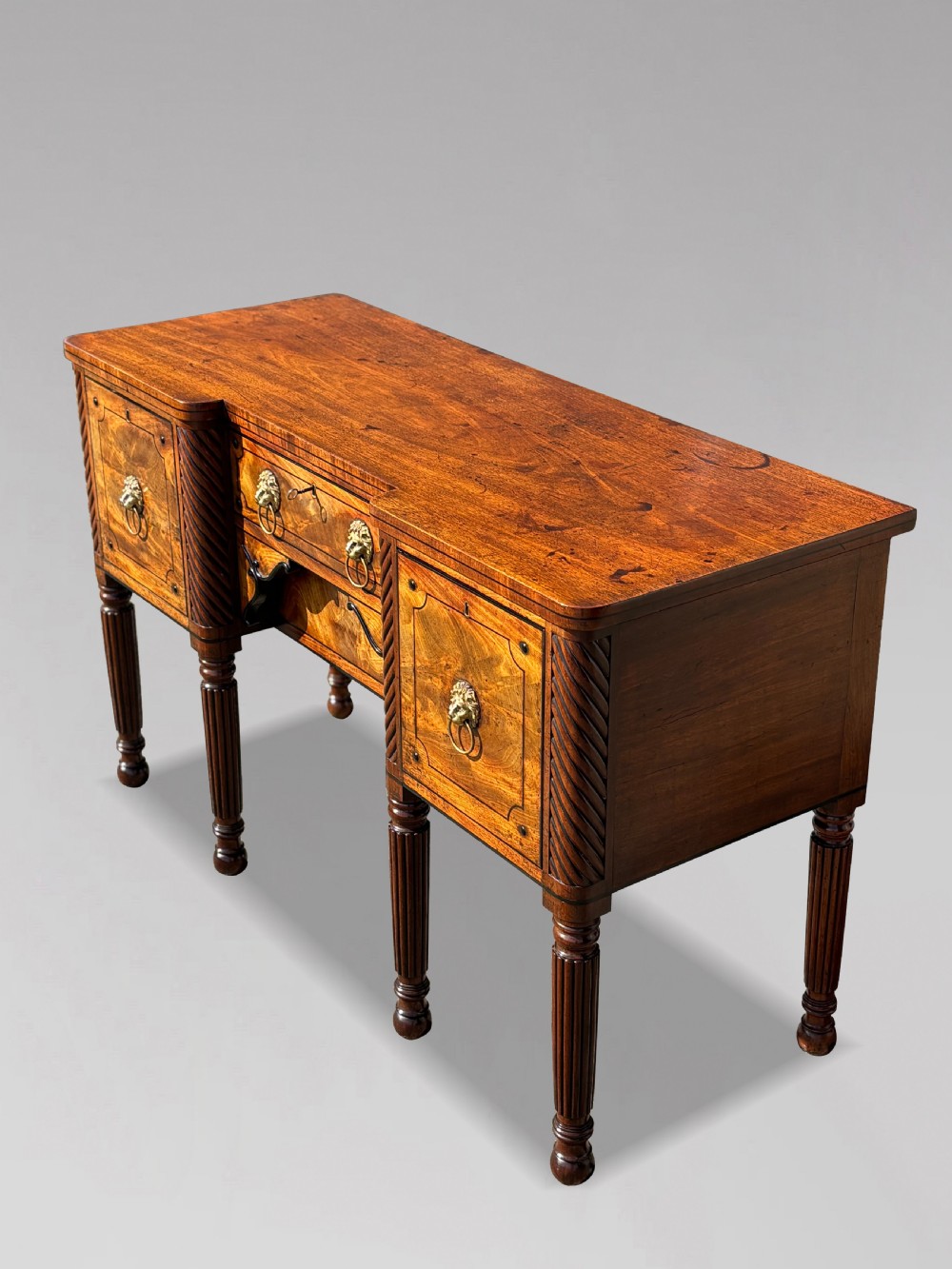 A 19th-century George IV flame mahogany breakfront sideboard featuring ebony stringing, lion-mask brass handles, spiral pilasters, and fluted legs. View XV