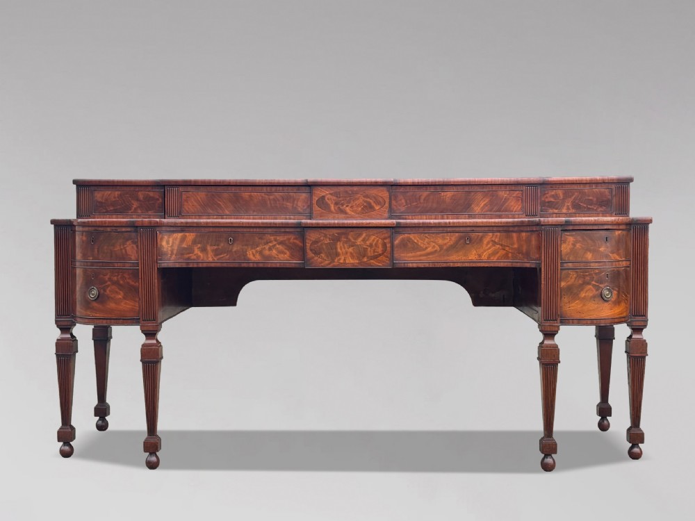 An 18th-century George III flame mahogany sideboard featuring a raised stage back, oval wood inlays, fluted side columns, and six turned reeded legs.