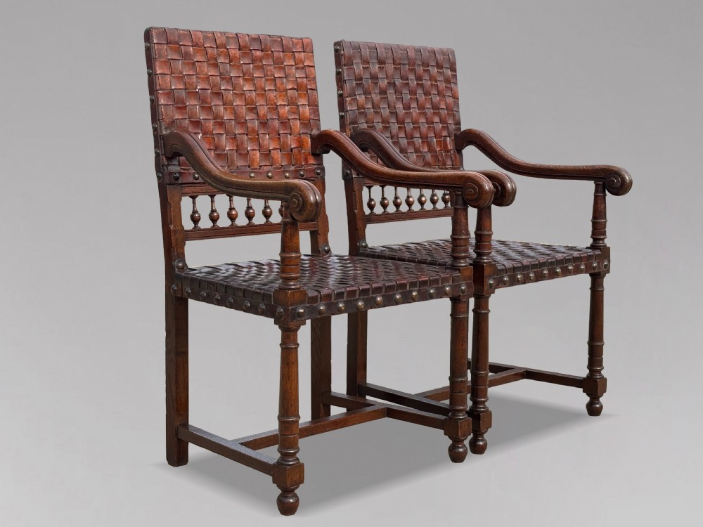 A pair of late 19th-century dark oak armchairs featuring woven brown leather backrests and seats with brass stud detailing and turned legs.