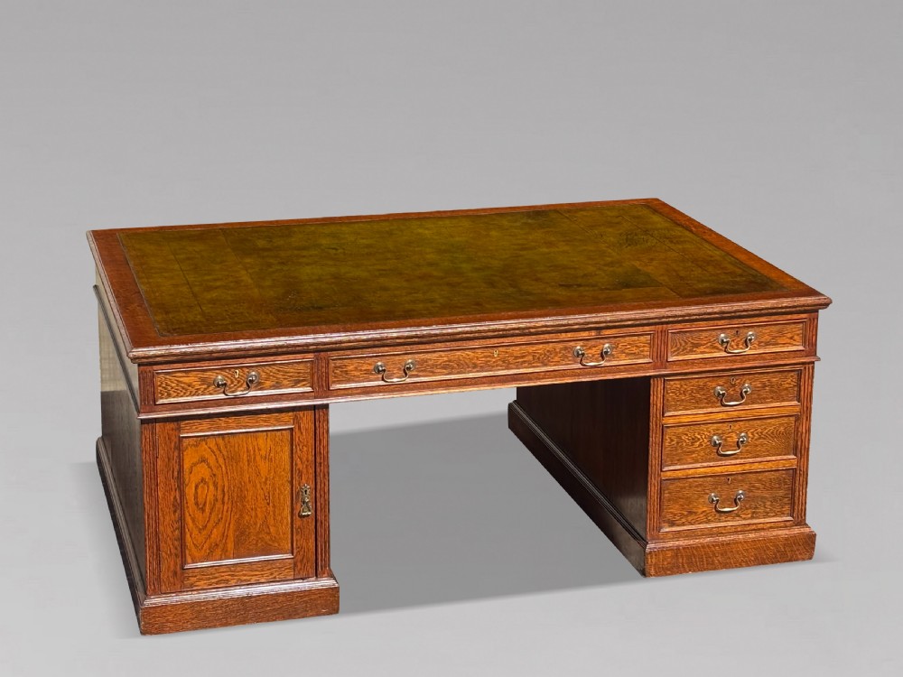 A late 19th-century oak partners desk featuring a green leather inset writing surface, twin pedestals with drawers and cupboards, and brass hardware.