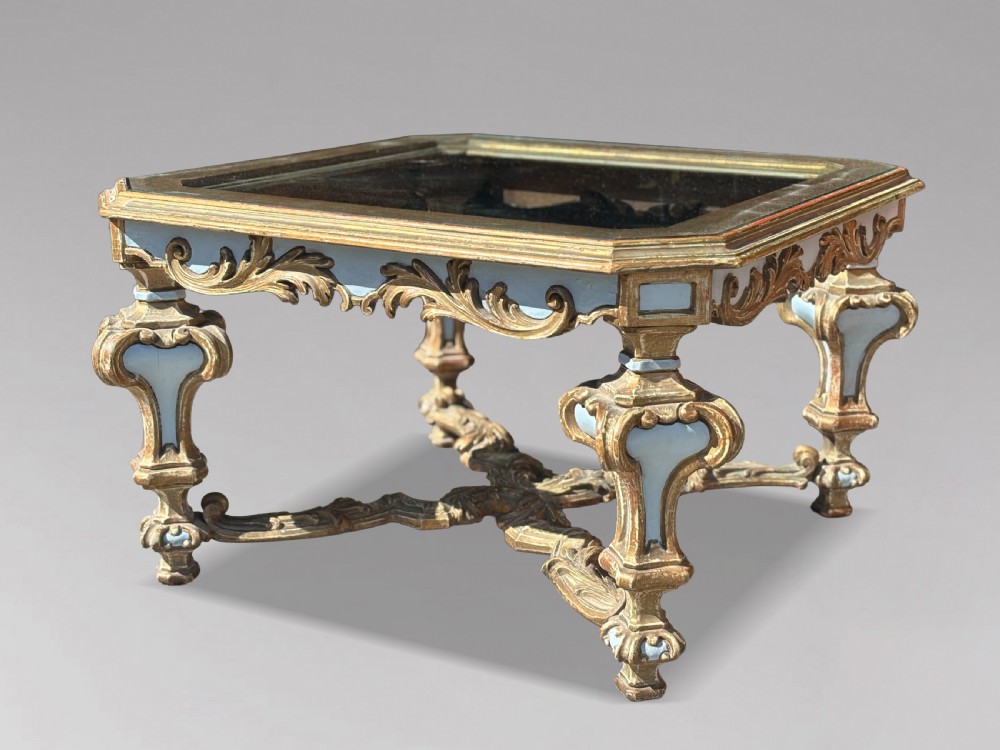 A square Italian Baroque style coffee table featuring a glass top, pale blue paint finish, carved gilt scrollwork frieze, ornate scrolled legs, and an X-shaped stretcher.