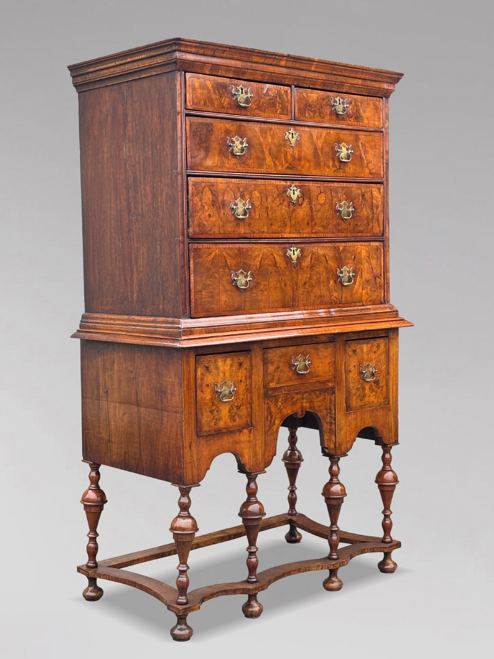 An 18th-century Queen Anne walnut chest on stand featuring figured veneers, brass drop handles, a shaped lower apron, and turned legs joined by stretchers.