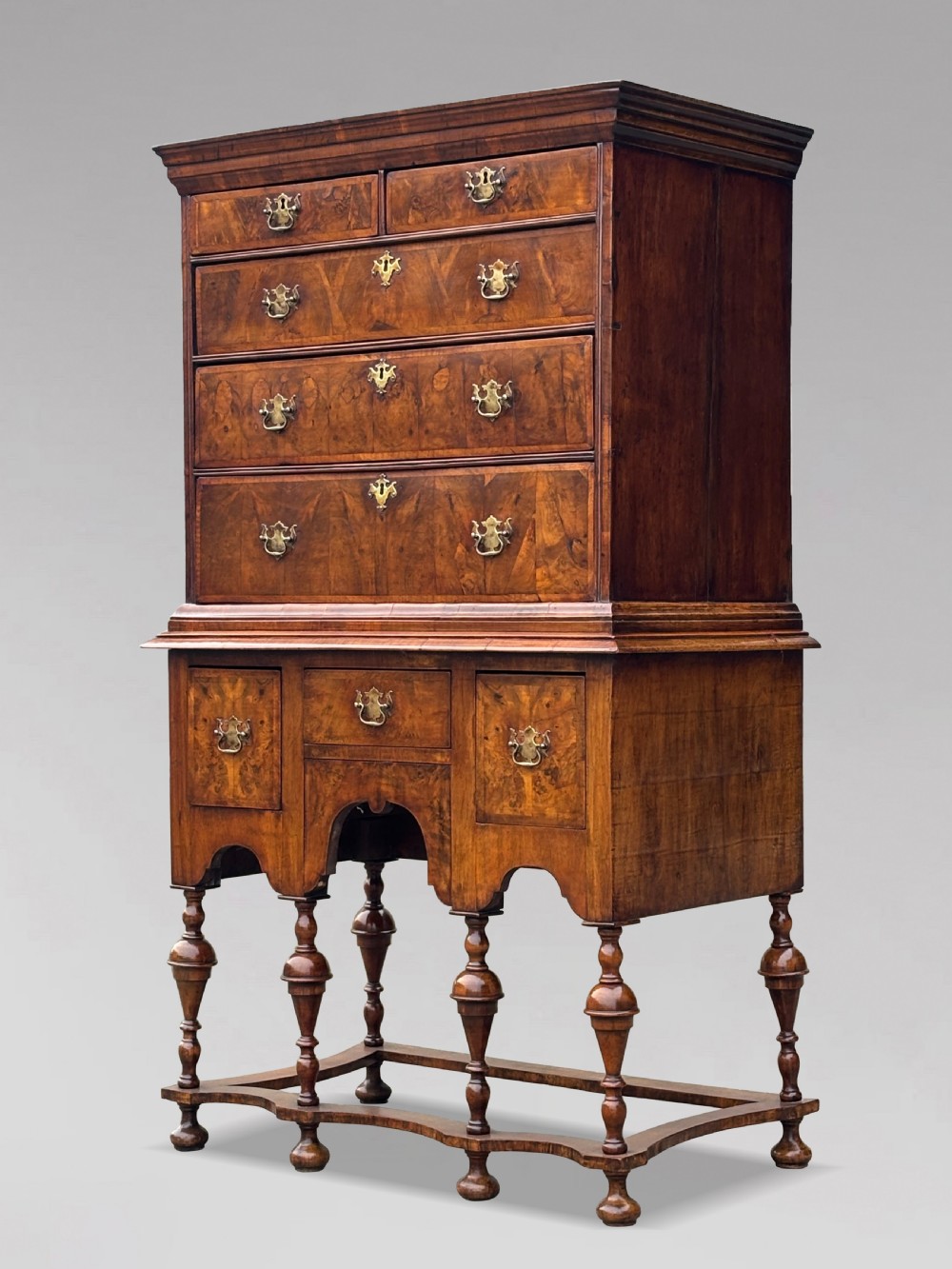 An 18th-century Queen Anne walnut chest on stand featuring figured veneers, brass drop handles, a shaped lower apron, and turned legs joined by stretchers. View V