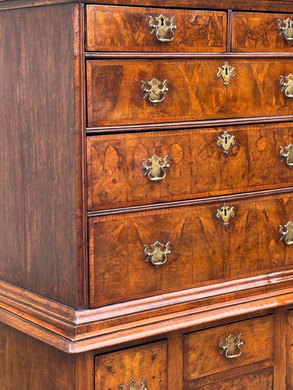 An 18th-century Queen Anne walnut chest on stand featuring figured veneers, brass drop handles, a shaped lower apron, and turned legs joined by stretchers. View X