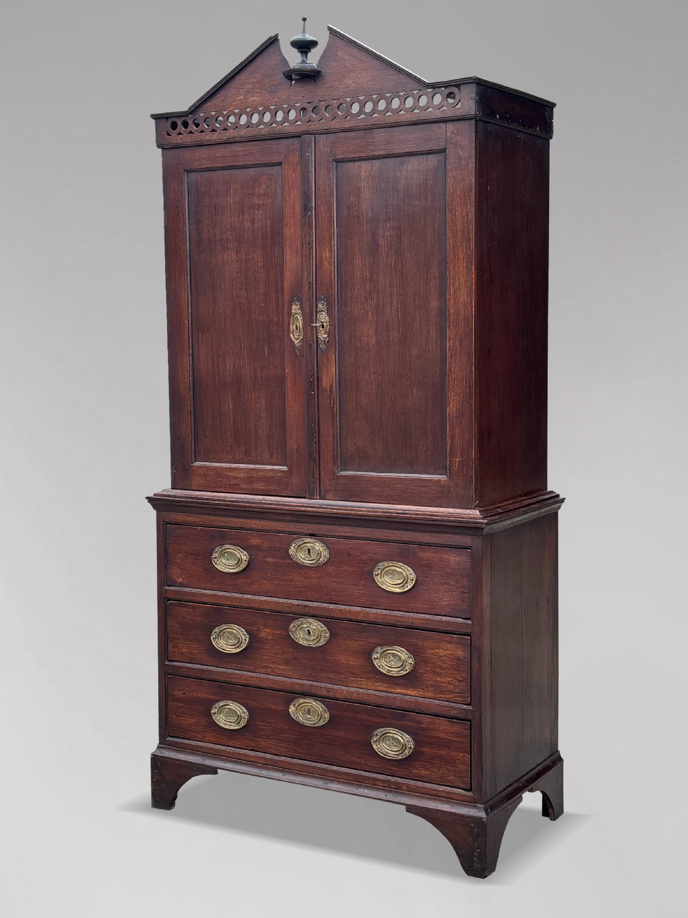 An 18th-century George III dark oak housekeepers cupboard featuring a fretwork gallery with a central turned finial, two upper panel doors, and three lower drawers with brass oval handles.