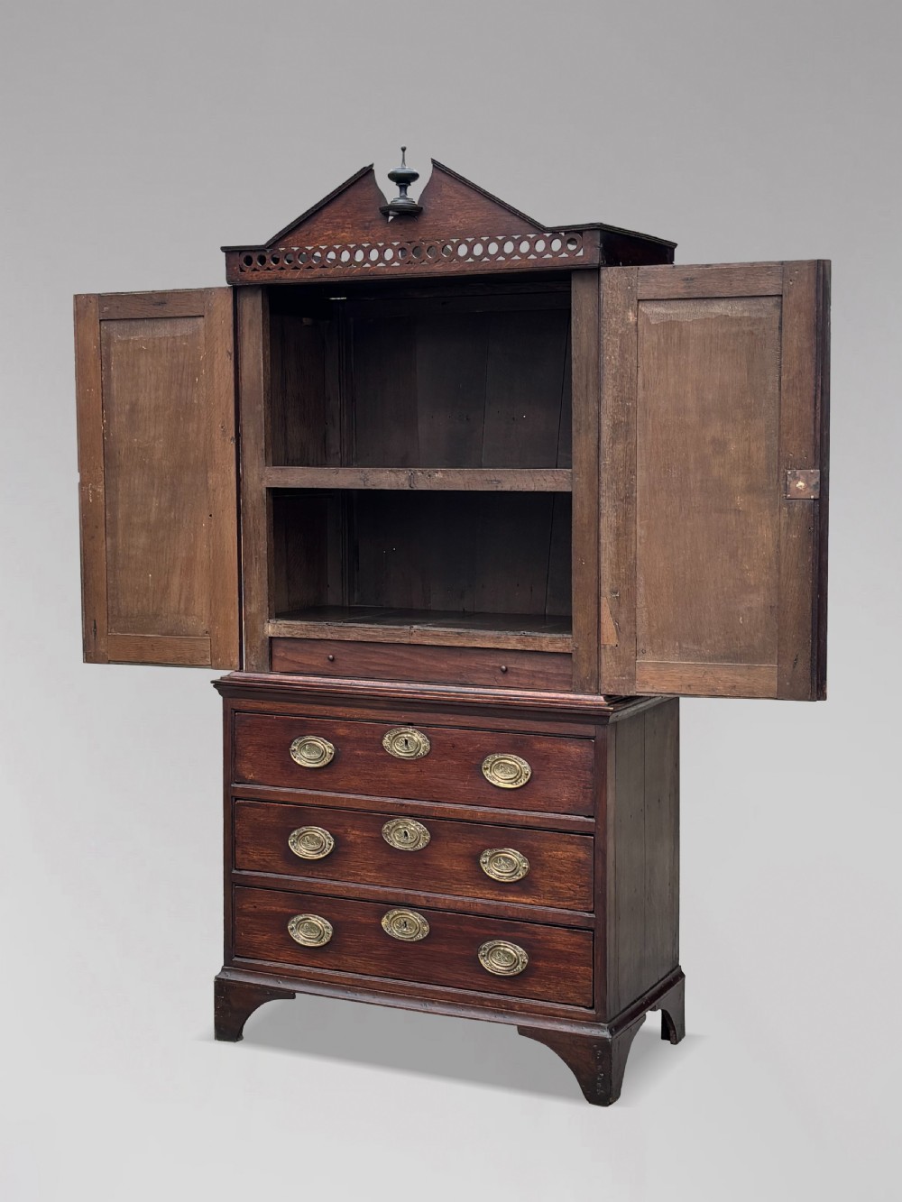 An 18th-century George III dark oak housekeepers cupboard featuring a fretwork gallery with a central turned finial, two upper panel doors, and three lower drawers with brass oval handles. View II