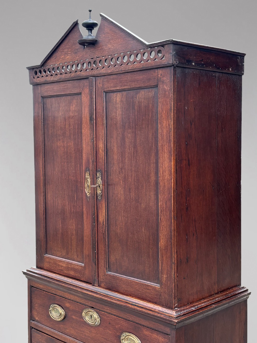 An 18th-century George III dark oak housekeepers cupboard featuring a fretwork gallery with a central turned finial, two upper panel doors, and three lower drawers with brass oval handles. View III