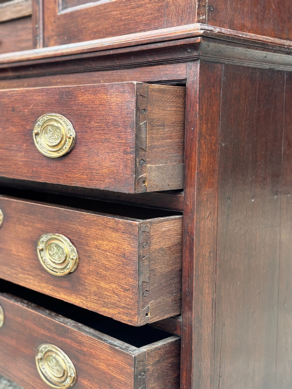 An 18th-century George III dark oak housekeepers cupboard featuring a fretwork gallery with a central turned finial, two upper panel doors, and three lower drawers with brass oval handles. View X