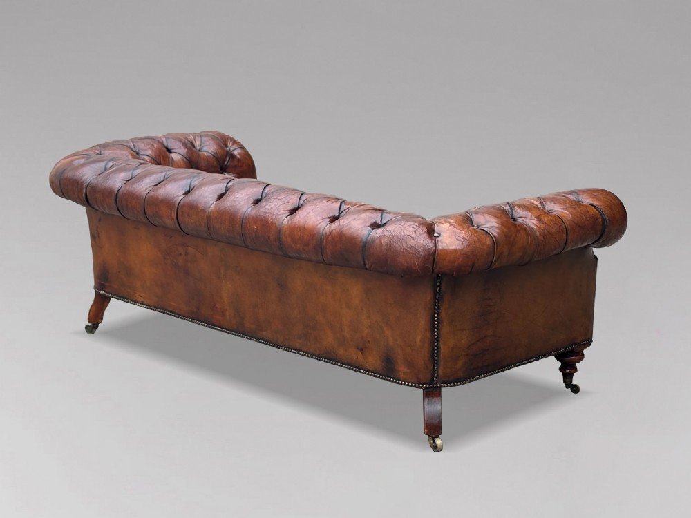 A late 19th-century Victorian brown leather Chesterfield sofa featuring deep buttoned upholstery, rolled arms, close studding, and turned legs with brass castors. View IV