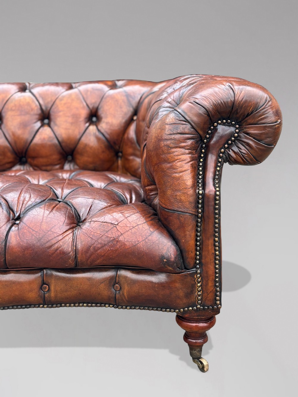 A late 19th-century Victorian brown leather Chesterfield sofa featuring deep buttoned upholstery, rolled arms, close studding, and turned legs with brass castors. View VI