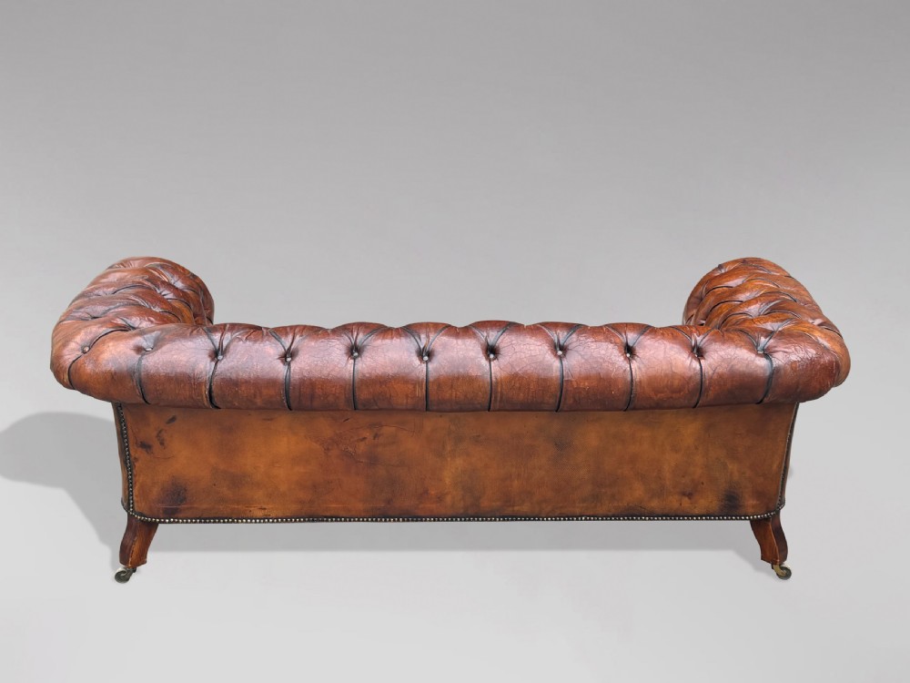 A late 19th-century Victorian brown leather Chesterfield sofa featuring deep buttoned upholstery, rolled arms, close studding, and turned legs with brass castors. View X